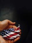 A close-up of two hands gently holding a small cutout of the United States map. The cutout is decorated with the American flag, displaying its stars and red-and-white stripes.