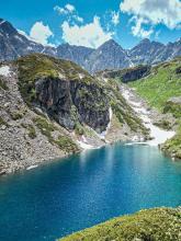 A mountain landscape featuring a serene turquoise alpine lake surrounded by rocky slopes and green hillsides